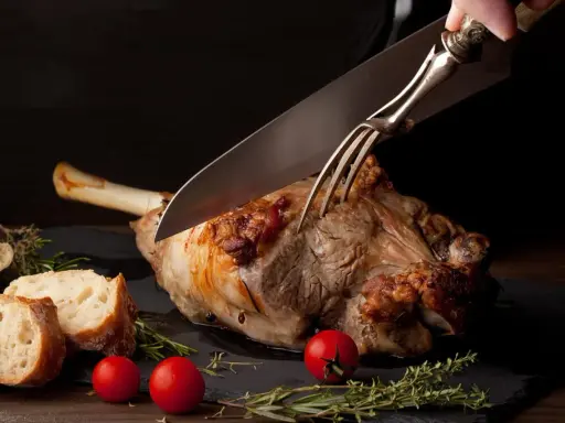 Une main utilise un couteau et une fourchette pour découper un gigot d'agneau rôti, prêt à être servi avec du pain et des tomates.