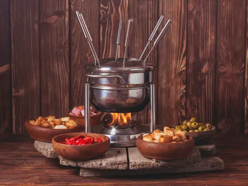 Un appareil à fondue bourguignonne chauffe, entouré de bols de croûtons, d'olives, de tomates séchées et de dés de viande. Idéal pour savoir que faire avec de la viande à fondue bourguignonne.