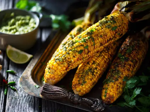 Épis de maïs grillés, parsemés d'herbes fraîches, servis sur un plateau avec une sauce verte et des quartiers de citron vert. Un délice parfait pour un repas mais au barbecue.
