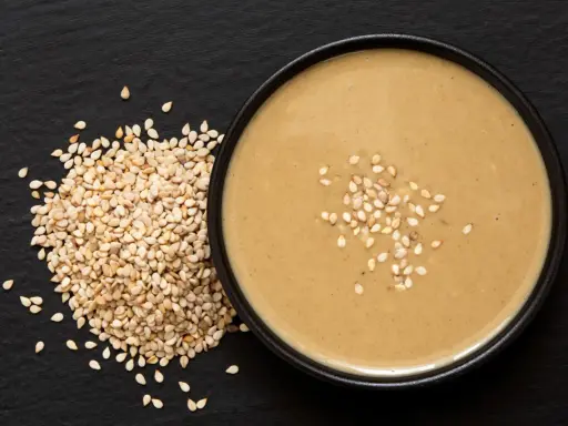 Un bol de sauce tahini onctueuse, garni de graines de sésame, à côté d'un tas de graines de sésame sur fond noir.