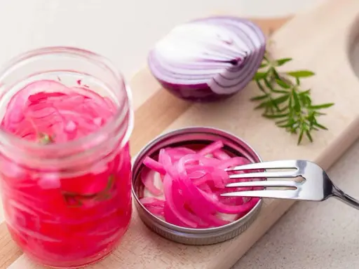 Pickles oignon rouge vibrants dans un bocal, avec une moitié d'oignon rouge et une branche de romarin sur une planche à découper.