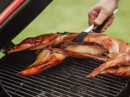 Un lapin entier est cuit sur un barbecue, sa peau dorée et croustillante. Une spatule retourne délicatement la viande pour une cuisson parfaite.