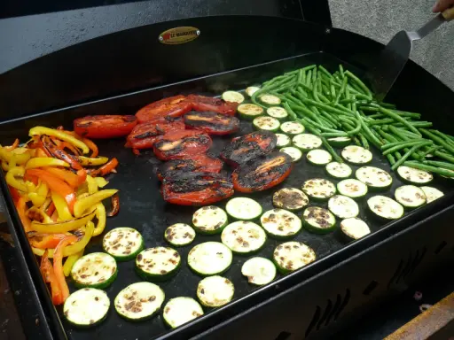Légumes variés (poivrons, courgettes, tomates, haricots verts) en cours de cuisson sur une plancha. Le temps de cuisson des légumes à la plancha est idéal pour un repas sain et savoureux.