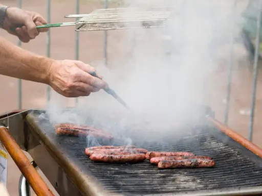 Des merguez grillent sur un barbecue, dégageant une fumée épaisse. Un homme les retourne avec une pince, se demandant s'il faut piquer les merguez pour une cuisson parfaite.