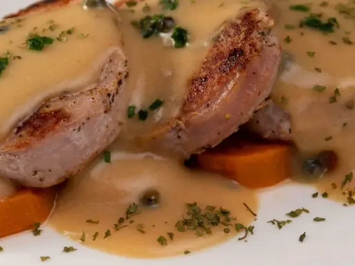 Tranches de gigot d'agneau nappées d'une sauce onctueuse au fond de veau, parsemées de persil et de câpres, sur un lit de carottes.