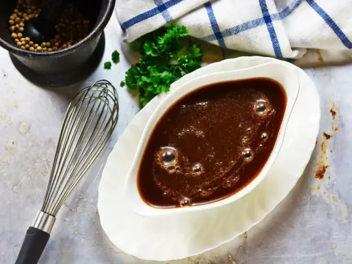 Sauce à la viande fumante dans un saucier, avec un fouet, du persil et un mortier rempli de grains de poivre.