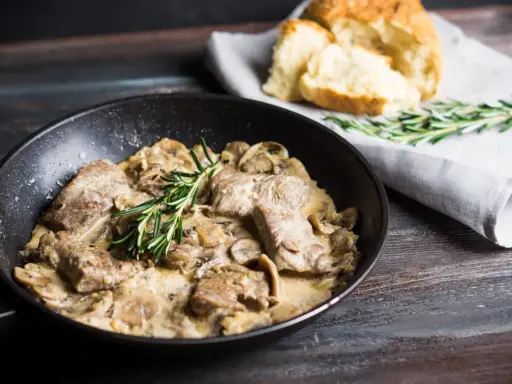 Délicieux sauté de porc crémeux aux champignons, garni de romarin frais, servi dans une poêle avec du pain croustillant à côté.