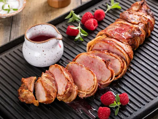 Filet mignon enrobé de bacon, tranché, servi avec des framboises et une sauce rouge sur une plaque de cuisson.