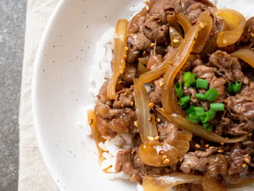 Un plat de riz blanc garni de fines tranches de bœuf aux oignons caramélisés, parsemé de graines de sésame et de ciboule ciselée. Un délice de boeuf aux oignons.