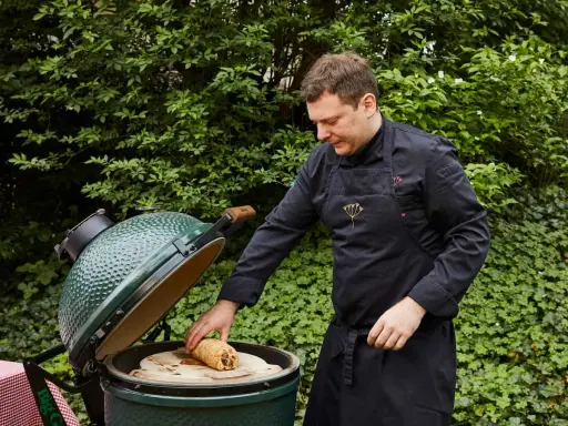 Un chef prépare un rôti sur une pierre à pizza dans un barbecue binchotan vert, entouré de verdure luxuriante.