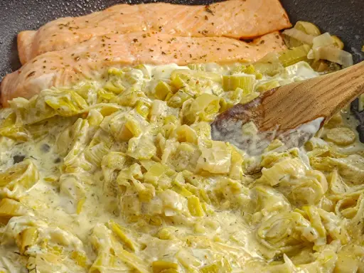 Filets de saumon poêlés sur une fondue de poireaux crémeuse, parsemée d'herbes. Une cuillère en bois remue ce plat savoureux.