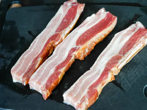 Trois tranches de lard fumé, prêtes pour une recette savoureuse, reposent sur une planche à découper noire.