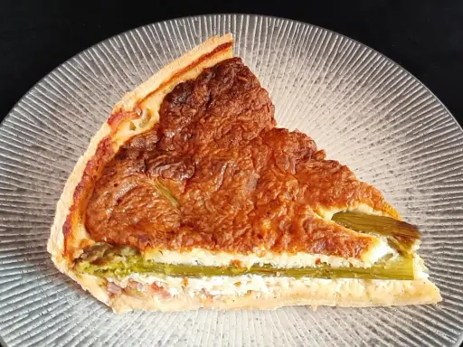 Tranche de tarte aux asperges blanches, garnie d'une préparation crémeuse et dorée, sur une assiette texturée.