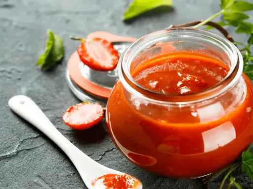 Un pot de ketchup maison, garni de fraises et d'une cuillère blanche, sur fond sombre.