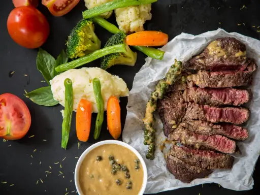 Un steak tranché, servi avec des légumes et deux types de sauces. L'une est une sauce crémeuse aux grains de poivre vert, l'autre est une sauce plus claire dans un bol.