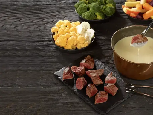 Un morceau de viande est trempé dans un caquelon de fondue. Des morceaux de viande, du brocoli, du chou-fleur et des légumes racines sont disposés sur une table en bois sombre.