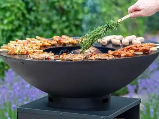 Un brasero moderne cuit brochettes et saucisses. On y voit comment fonctionne un brasero, avec une branche de romarin pour parfumer.