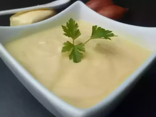 Un bol de beurre blanc crémeux, garni d'une feuille de persil. Des échalotes et un morceau de pain sont visibles en arrière-plan.