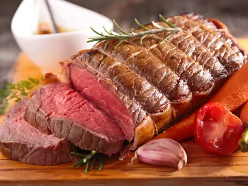 Un délicieux roti de boeuf saignant, garni de romarin, servi avec des carottes, une tomate et de l'ail sur une planche en bois.