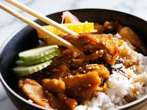 Un bol de riz garni de poulet teriyaki, de concombre, d'omelette et de gingembre mariné, prêt à être dégusté avec des baguettes.