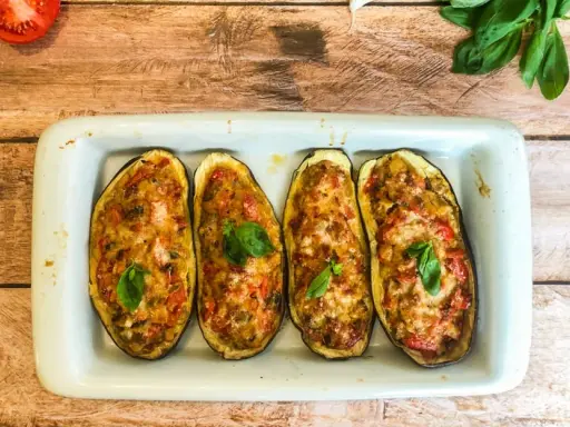 Quatre moitiés d'aubergines garnies de légumes et de fromage gratiné, décorées de feuilles de basilic frais.
