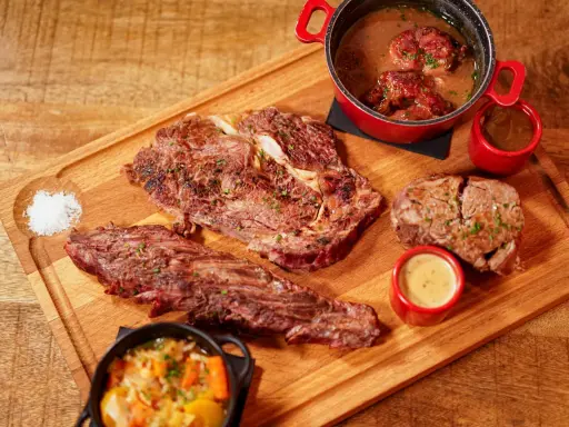 Assortiment de viande à griller, steak, et ragoût de bœuf, servi avec des sauces et des légumes sur une planche en bois.