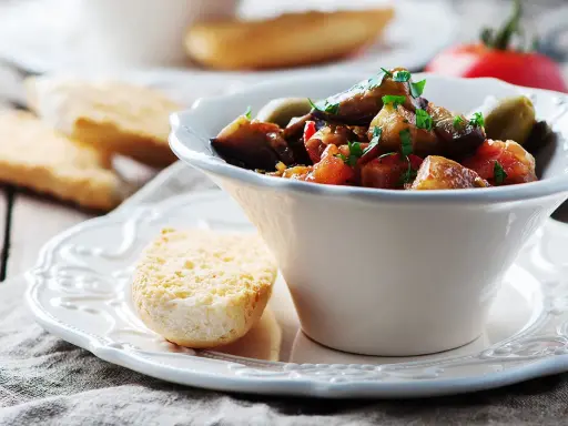 Un bol de caponata sicilienne, plat sicilien aux aubergines, tomates et olives, garni de persil frais. Servi avec du pain grillé.