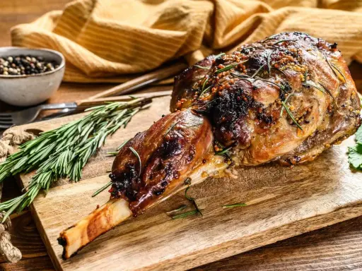 Un gigot d'agneau tendre, rôti à la perfection, est présenté sur une planche en bois avec du romarin frais et des grains de poivre.