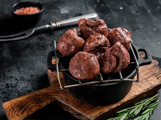 Un plat de rognons de veau grillés, servis dans une cocotte en fonte sur une planche en bois, avec une pincée de sel rose et une branche de romarin.