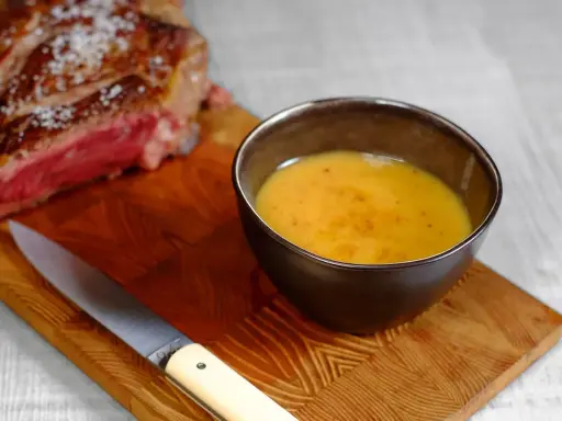 Un bol de sauce pour côte de bœuf, onctueuse et dorée, à côté d'une côte de bœuf saignante et d'un couteau sur une planche en bois.
