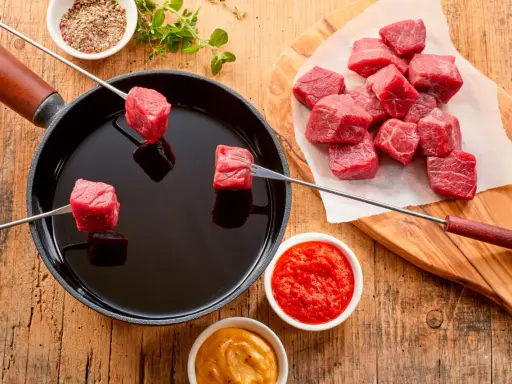 Préparation d'une fondue chinoise : des cubes de viande rouge sur des brochettes trempent dans un bain d'huile chaude. Quel accompagnement fondue viande ? Sauces et épices sont à portée de main.