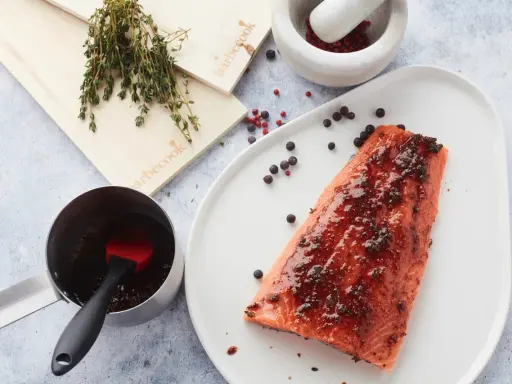 Un filet de saumon mariné est prêt à être cuit sur une planche de cèdre. Des herbes fraîches et des grains de poivre complètent la scène.