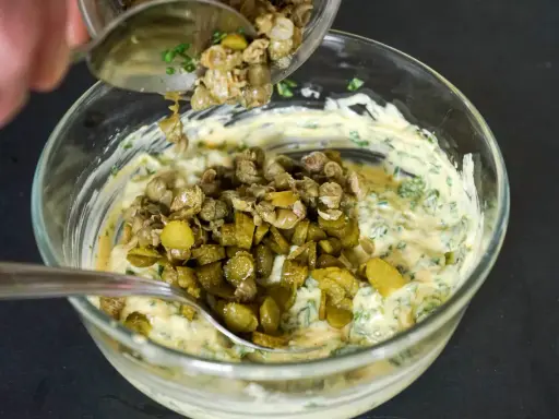 Ajout de câpres et cornichons hachés à une base crémeuse pour préparer une sauce gribiche savoureuse.