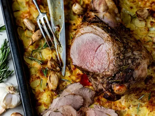 Un gigot d'agneau rôti, tranché, servi sur un lit de pommes de terre gratinées avec de l'ail et du romarin.