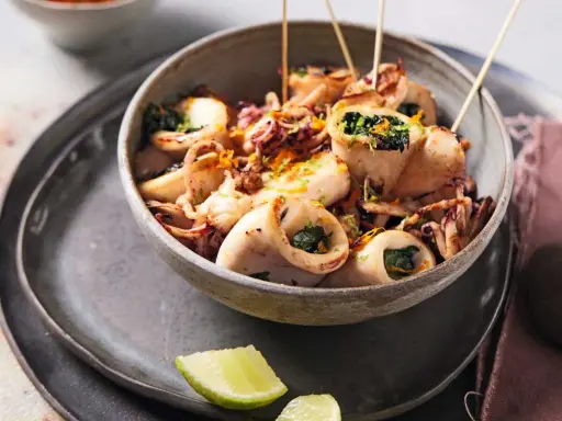 Calamars à la plancha farcis aux herbes, servis dans un bol avec des zestes d'orange et des quartiers de citron vert.