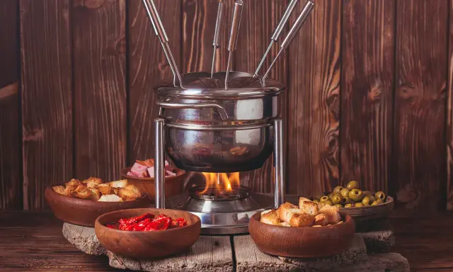 Un appareil à fondue bourguignonne chauffe, entouré de bols de croûtons, d'olives, de tomates séchées et de dés de viande. Idéal pour savoir que faire avec de la viande à fondue bourguignonne.
