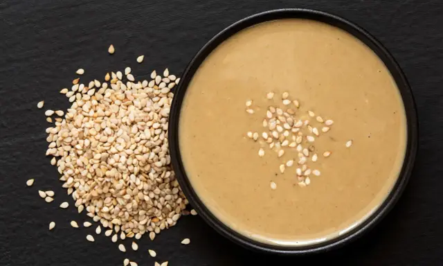 Un bol de sauce tahini onctueuse, garni de graines de sésame, à côté d'un tas de graines de sésame sur fond noir.