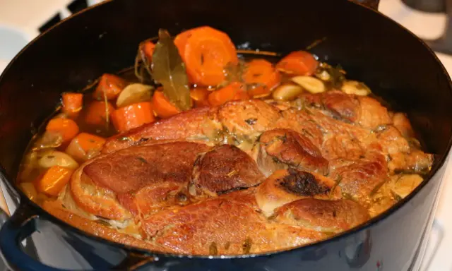 Rouelle de porc mijotant dans une cocotte avec des carottes, de l'ail et des herbes.