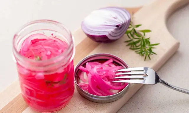 Pickles oignon rouge vibrants dans un bocal, avec une moitié d'oignon rouge et une branche de romarin sur une planche à découper.