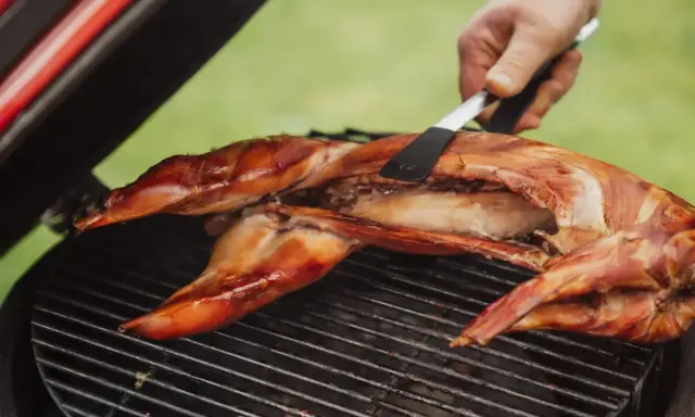 Un lapin entier est cuit sur un barbecue, sa peau dorée et croustillante. Une spatule retourne délicatement la viande pour une cuisson parfaite.