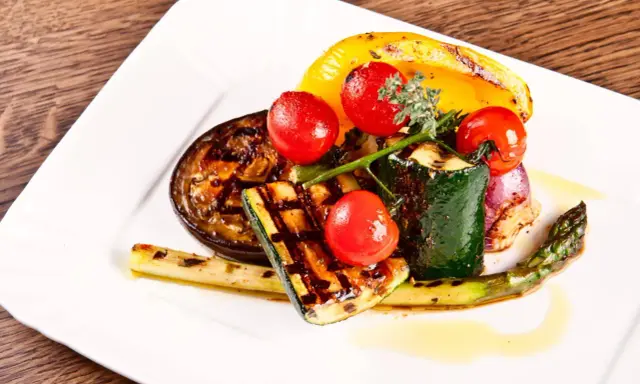 Légumes grillés : courgette, tomate, poivron jaune, aubergine, asperge et oignon rouge, arrosés d'une huile dorée.