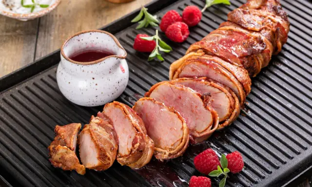 Filet mignon enrobé de bacon, tranché, servi avec des framboises et une sauce rouge sur une plaque de cuisson.