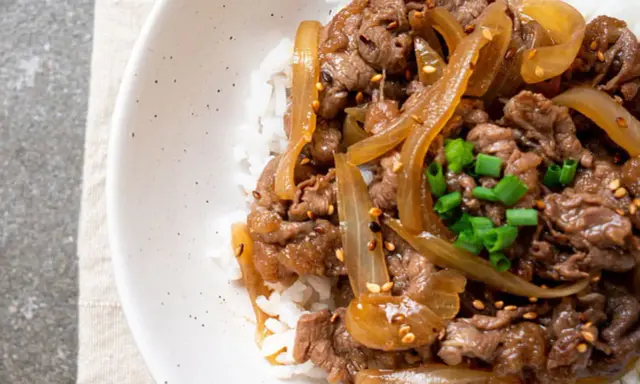 Un plat de riz blanc garni de fines tranches de bœuf aux oignons caramélisés, parsemé de graines de sésame et de ciboule ciselée. Un délice de boeuf aux oignons.