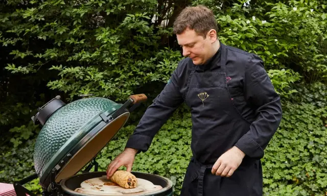 Un chef prépare un rôti sur une pierre à pizza dans un barbecue binchotan vert, entouré de verdure luxuriante.