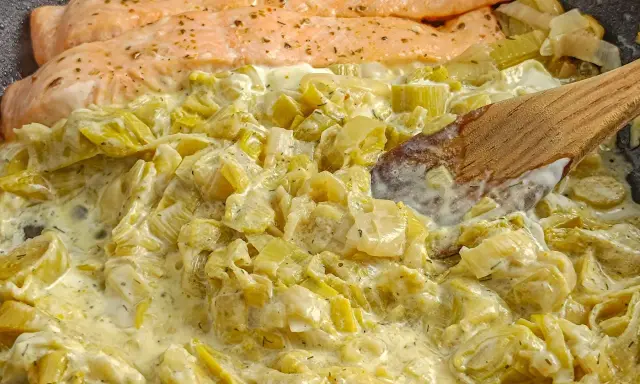 Filets de saumon poêlés sur une fondue de poireaux crémeuse, parsemée d'herbes. Une cuillère en bois remue ce plat savoureux.