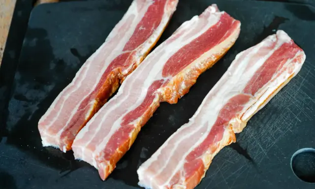 Trois tranches de lard fumé, prêtes pour une recette savoureuse, reposent sur une planche à découper noire.