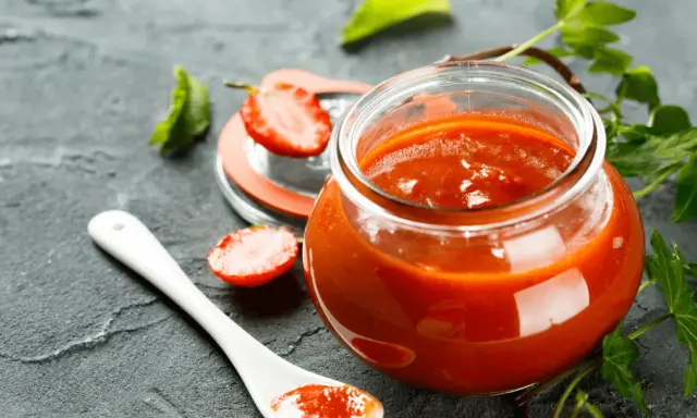 Un pot de ketchup maison, garni de fraises et d'une cuillère blanche, sur fond sombre.