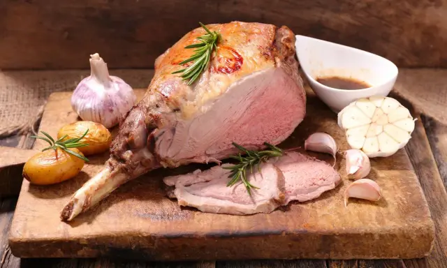 Un gigot d'agneau rôti, tendre et juteux, servi avec des pommes de terre, de l'ail et du romarin sur une planche en bois.