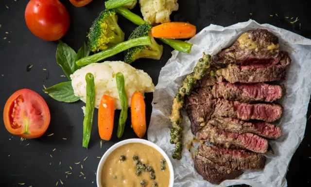 Un steak tranché, servi avec des légumes et deux types de sauces. L'une est une sauce crémeuse aux grains de poivre vert, l'autre est une sauce plus claire dans un bol.