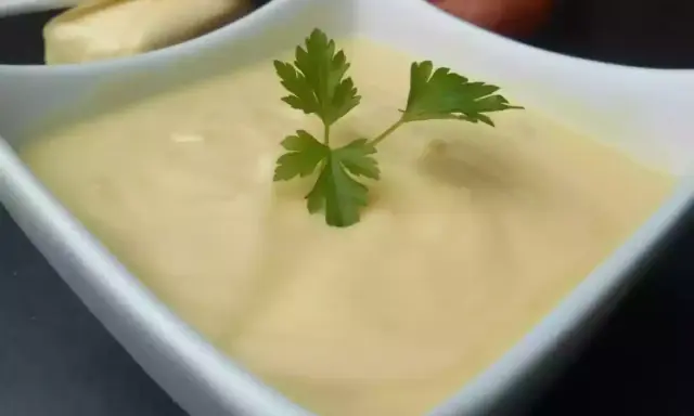 Un bol de beurre blanc crémeux, garni d'une feuille de persil. Des échalotes et un morceau de pain sont visibles en arrière-plan.